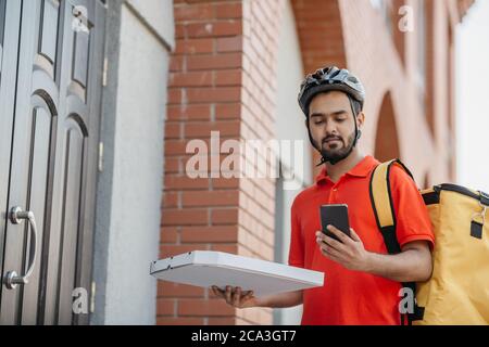 Schnelle Lieferung von Pizza per Kurier in der Stadt. Serious Mann in Helm und Tasche, hält Karton mit Lebensmitteln und Uhren auf dem Smartphone Stockfoto