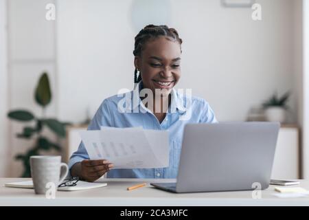 Papierkram. Fröhliche schwarze Millennial Geschäftsfrau arbeiten mit Laptop und Dokumente im Büro Stockfoto