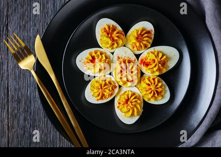 Klassische deviled Eier gefüllt mit Senf, Mayonnaise, Weißessig bestreut mit geräuchertem Paprika auf schwarzem Teller mit goldenem Besteck auf einem dar hölzernen b Stockfoto