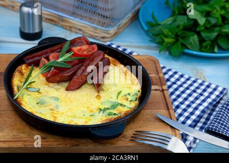 Omelette mit Wurst und Tomaten in einer Pfanne auf dem Tisch. Appetitliches Frühstück. Ein Zweig Rosmarin, ein hölzernes Gewürz. Jagt Würstchen. Leckere Speisen gebraten Stockfoto