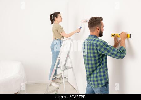 Content junge Frau auf der Trittleiter und Malerei Wand, während ihr Freund Zeichnung Linie mit Ebene Stockfoto