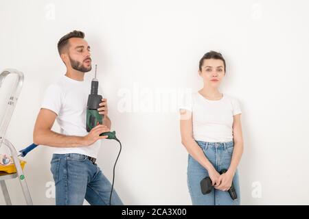 Cool junge Reparaturman weht an Bohrer nach der Herstellung von Löchern, während seine Freundin mit Akku-Schraubendreher Gefühl Erschöpfung Stockfoto