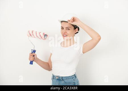 Portrait von positiven selbstbewussten jungen Frau Anpassung Hardhat und halten Farbe Rolle gegen weiße Wand Stockfoto