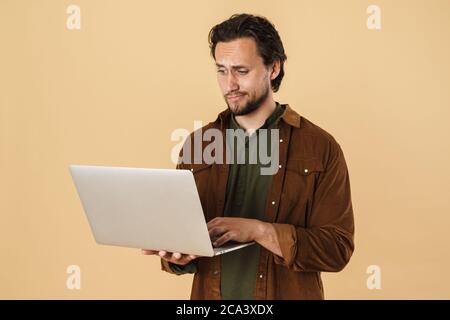 Bild von verwirrt bärtigen Mann in Casual Shirt tippen auf Notebook isoliert über beige Hintergrund Stockfoto