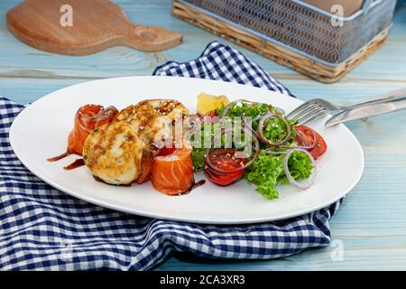 Fischkuchen mit Salat, Tomaten und roten Zwiebeln in einem Restaurant auf einem weißen Teller. Roter Lachsfisch. Fischmenü. Nahaufnahme. Gesunde Omega-3-Lebensmittel auf blau Stockfoto