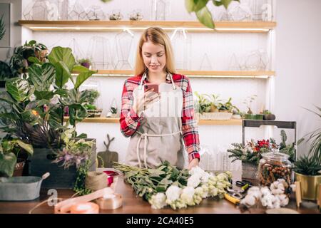 Schöne Floristin mit blonden Haaren bei der Arbeit, im Gespräch mit Freund und Lächeln, haben Freizeit bei der Arbeit tragen Schürze, isoliert in hellen Raum umgeben Stockfoto