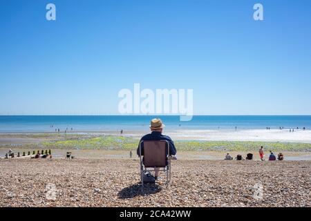 Küstenansicht mit einer männlichen Einzelfigur, die allein auf dem Campingstuhl unter blauem Himmel sitzt und die Menschen den Sommer am Strand während der Absperrung in Wort genießen Stockfoto