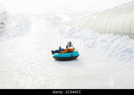 Kleiner Junge rollt auf der Eisrutsche auf dem Schlauch herunter. Winterzeit. Spaß und Spiele, Outdoor-Aktivitäten-Konzept. Stockfoto