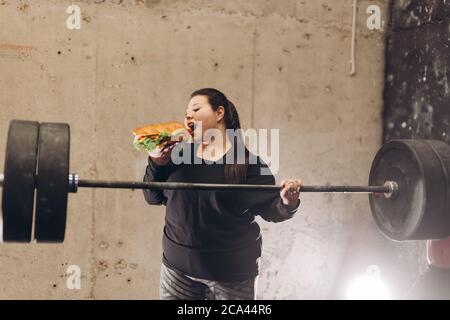 Fettleibige junge starke Frau arbeitet mit Langhantel in der Turnhalle. Nahaufnahme Foto. Essen Mädchen während des Trainings. Frau ist wütend auf Hamburger. Stockfoto