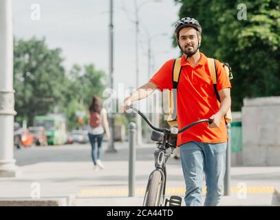 Ampel und Fußgängerüberweg in der Stadt. Freundlicher Kurier im Helm mit großer gelber Tasche und Fahrrad Stockfoto