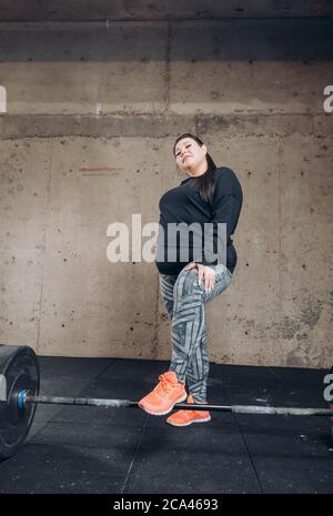 Angenehme übergewichtige Mädchen stehen im Fitnessstudio und lehnte sich auf die Langhantel. Foto in voller Länge. Rest Stockfoto