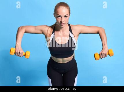 Motivierte Sportlerin macht Übung für Arme. Weibliche Modell mit Hanteln auf blauem Hintergrund ausarbeiten. Stärke und Motivation. Isolierter blauer Rücken Stockfoto