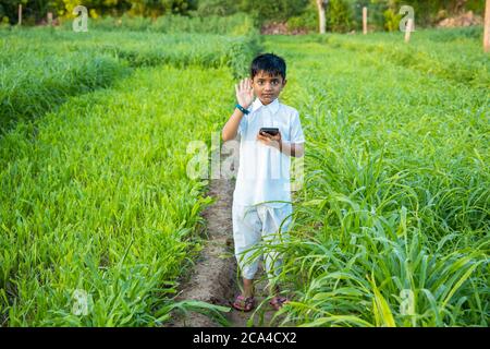 Portrait auf niedlichen indischen Kind mit Smartphone winkende Hand stehen in der Landwirtschaft grünes Feld, Technologie im ländlichen indien Konzept. Stockfoto