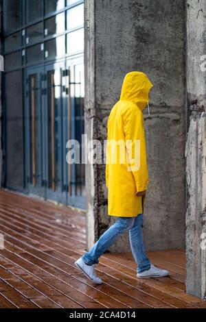 Mann in gelber Regenjacke mit Kapuze auf Spaziergängen bei Regenwetter in der Nähe des Gebäudes auf nassem Holzboden, Seitenansicht. Im Freien. Stockfoto