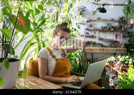 Frau Gärtnerin in Brille tragen orange Jeans Overalls, sitzen auf Stuhl im Gewächshaus, arbeiten auf Laptop an einem Projekt von Pflanzen umgeben. Home gar nicht Stockfoto