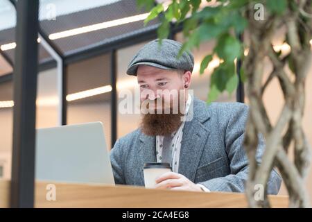 Portrait eines brutalen bärtigen Hipsters mit Wollblazer, Mütze auf Laptop sitzend im Café/Restaurant, Kaffee aus einer Papiertasse trinkend, Indoo Stockfoto
