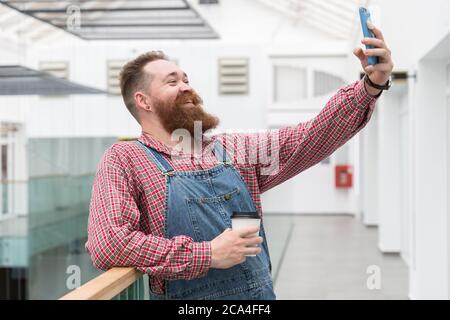 Lächelnder bärtiger Barbier in blauen Overalls, kariertes Hemd, Kaffee aus einer Papiertasse trinken, bei einem Videoanruf sprechen oder ein Selfie auf Smartph machen Stockfoto