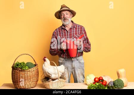 Fröhlicher alter Mann kam gerade aus seinem Garten, wo er Pflanzen gewässert, steht mit Gießkanne, brachte er üppiges Gemüse und Eier zu Hause für Frau, g Stockfoto