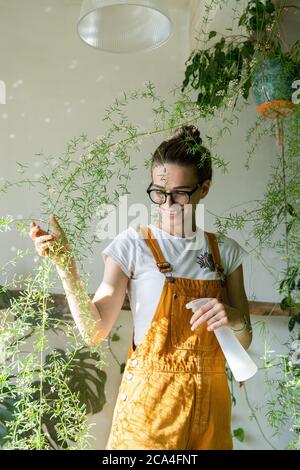 Lächelnde junge Frau Gärtnerin in orange Overalls sprühen üppige Spargelfarn Zimmerpflanze in ihrem Blumengeschäft. Viel Grün zu Hause. Liebe zu Pflanzen. Innen Stockfoto