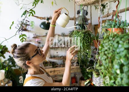Frau Gärtner in orange Overalls Bewässerung Topfpflanze im grünen Haus von hängenden Pflanzen umgeben, mit weißen Gießkanne Metall. Home Gardeni Stockfoto