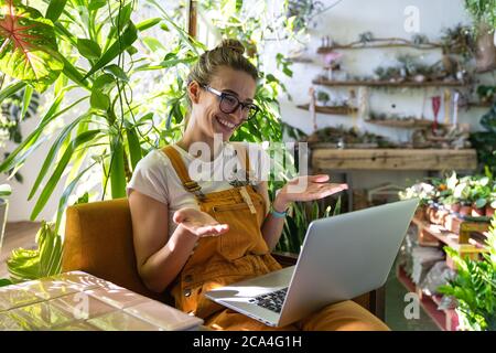 Frau Gärtnerin in Brille tragen orange Overalls, sitzen auf einem Stuhl im grünen Haus, mit Laptop nach der Arbeit, lächeln und sprechen auf Video-Anruf surrounde Stockfoto