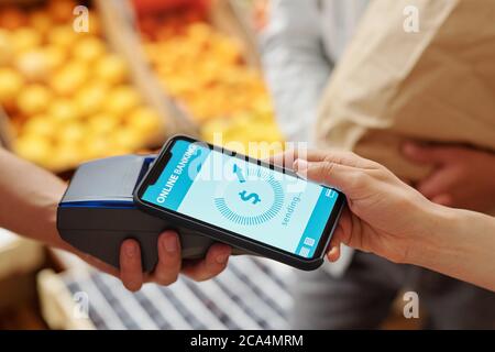 Nahaufnahme einer unkenntlichen Frau, die ihr Smartphone auf den Terminal legt, während sie auf dem Lebensmittelmarkt online für Produkte bezahlt Stockfoto