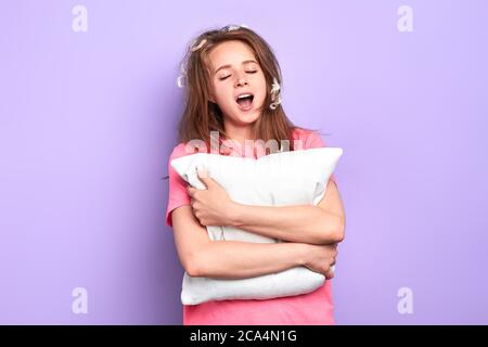 Vorderansicht eines hübschen Teenagers, das nach einem guten Schlaf gähnt, mit geschlossenen Augen und Federn im Haar steht, Kissen hält, sich entspannt fühlt, bereit dazu Stockfoto