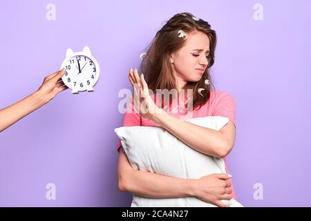 Studio Porträt von unzufriedenen jungen Dame hält weiße Kissen, Wellen beiseite Wecker, unglücklich, nicht genug Schlaf zu haben, müde, früh aufzuwachen, brauchen Stockfoto