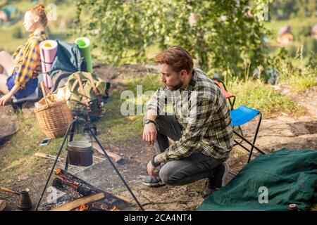 Abenteuer, Reisen, Tourismus und Menschen Konzept -junge gutaussehende Mann sitzt neben Lagerfeuer in Camping, volle Länge Seitenansicht Foto. Copy space.relaxati Stockfoto