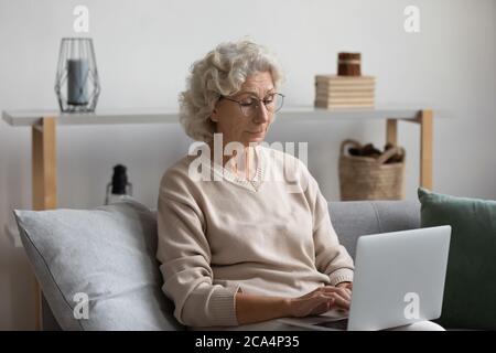 Ältere Frau in einer Brille, die auf einem Laptop auf der Couch sitzt Stockfoto