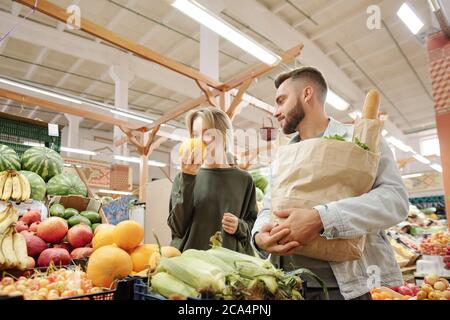 Junge Frau riecht Wassermelone bei der Wahl es auf Bauernmarkt, ihr Freund hält volle Papiertüte Stockfoto
