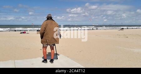 Der alte Mann mit Mantel, kurzer Hose, Hut und Spazierstock blickt auf das Meer an einem Strand in Holland Stockfoto