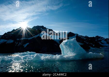 Hintergrundbeleuchteter Eisberg mit Sonnenstarburst über schneebedeckten Berg als Hintergrund zeigt Mangel an Eisschollen in Fjord aufgrund der globalen Erwärmung Klimakrise. Stockfoto