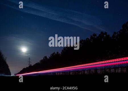 Leichte Spuren von Fahrzeugen, die auf einer ländlichen Straße fahren, umgeben von zwei Reihen von Bäumen, spät in der Nacht mit Mond am Himmel. Stockfoto