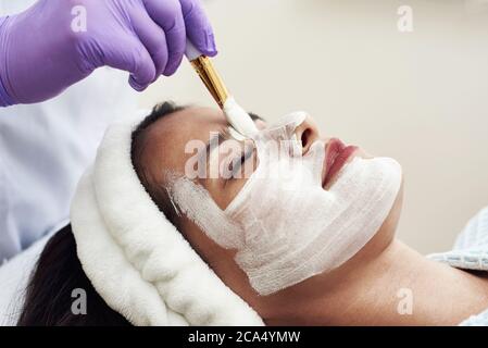 Eine ausgezeichnete Kosmetikerin trägt mit einem Pinsel eine weiße Anti-Aging-Feuchtigkeitsmaske auf das Gesicht auf. Stockfoto