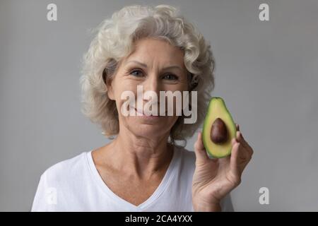 Lächelnde ältere Frau, die die Kamera mit der Hälfte der Avocado betrachtet Stockfoto