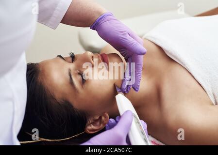 Eine Frau macht eine Ultraschallreinigung des Gesichts und der Haut zu einem Klienten. Moderne Ausstattung. Junge hübsche Frau, die Behandlungen in Schönheitssalons erhält und auf der Couch liegt. Stockfoto