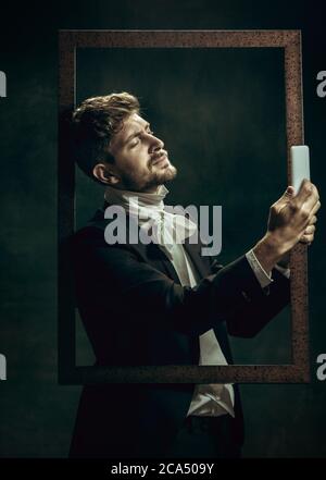 Selfie wird aufgenommen. Junger Mann in Anzug als Dorian Gray isoliert auf dunkelgrünem Hintergrund. Retro-Stil, Vergleich von Epochen Konzept. Schöne männliche Modell wie klassische Literatur Charakter, altmodisch. Stockfoto