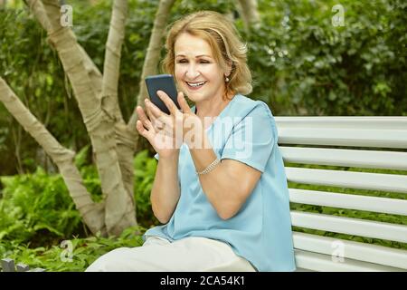 Aktive Rentnerin Attraktive weiße Frau etwa 60 Jahre alt mit Handy in den Händen sitzt auf der Bank im öffentlichen Park. Sie lächelt in ihrem Ga Stockfoto
