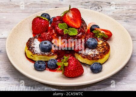 Hüttenkäse-Pfannkuchen mit frischen Beeren und Erdbeersoße. Gesundes Frühstückskonzept. Reife Heidelbeere, duftende Minzblätter. Holzbretter hintergr Stockfoto