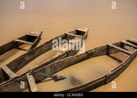 Drei alte Holzboote dockten an einem schmutzigen Fluss an. Eines der Boote hat zwei Schaufeln darin. Stockfoto