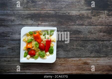 Frischer gesunder Salat mit Nudeln, Tomaten, Paprika, Käsewürfeln und Basilikum auf einem weißen quadratischen Teller. Dunkler Holzhintergrund. Stockfoto