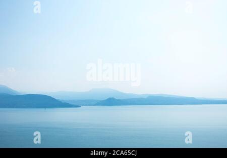 Blick auf den Gardasee am frühen Morgen oder Abend. Das Wasser ist ruhig, der Himmel teilweise bewölkt. Kleine Boote tanzen auf dem Wasser. Die Stimmung ist verträumt. Stockfoto