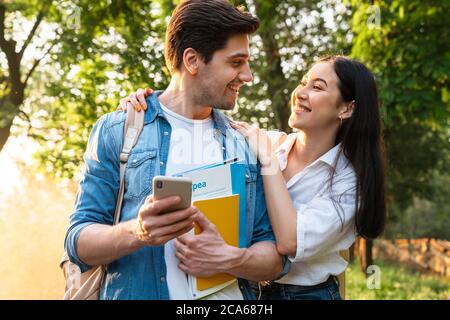 Bild eines fröhlichen multikulturellen Studentenpaares mit Handy und Lächeln beim Spaziergang im grünen Park Stockfoto