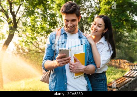 Bild eines fröhlichen multikulturellen Studentenpaares mit Handy und Lächeln beim Spaziergang im grünen Park Stockfoto