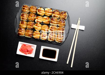 Sushi-Set serviert auf einem Steinschiefer auf dunklem Hintergrund. Sushi-Brötchen gebacken mit Sesamsamen, Unagi-Sauce, Ingwer und Essstäbchen. Sushi Rollen Foto für Stockfoto