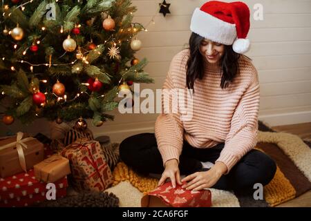 Stilvolle junge Frau Verpackung weihnachtsgeschenk in Papier unter weihnachtsbaum mit Lichtern in modernen Raum. Happy girl in santa Hut und Pullover Vorbereitung Stockfoto