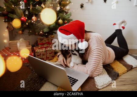 Junge Frau sitzt mit niedlichen Katze und Laptop auf dem Teppich unter weihnachtsbaum mit Geschenken. Happy girl in santa Hut küssen Kitty und arbeiten oder chatten Stockfoto