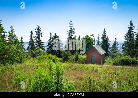 Ein kleines rotes Ziegelgebäude liegt in einem üppigen grünen Feld, umgeben von Bäumen mit einer Bucht von Wasser im Hintergrund. Stockfoto