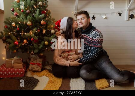 Frohe Weihnachten. Stilvolle glückliche Paar Austausch weihnachtsgeschenke unter weihnachtsbaum mit Lichtern. Junge Familie küssen und halten präsent, glückliche Mutter Stockfoto